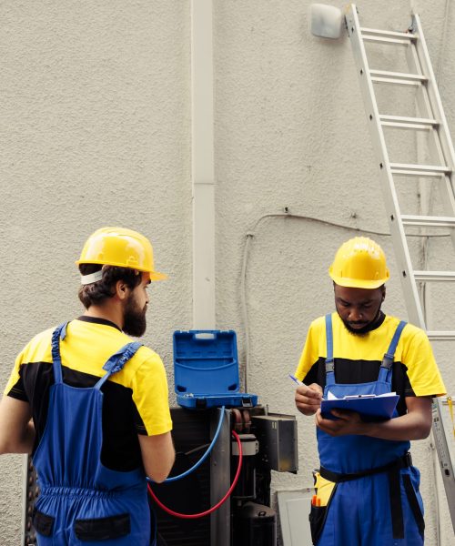 Professional worker using manifold indicators to read pressure of liquids and gases in hvac system while qualified electrician writes outdoor condenser inspection and safety check results on clipboard